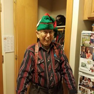 An elderly man wearing a colorful hat and striped shirt, smiling in his home.