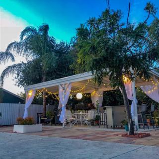 A cozy outdoor patio area with a pergola, draped curtains, and warm lighting surrounded by lush palm trees.