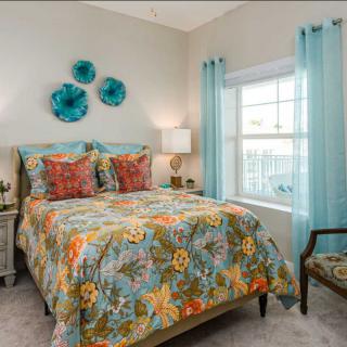 A vibrant bedroom with colorful bedding, decorative pillows, and wall art, enhanced by natural light from a large window.