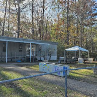 A cozy mobile home set in a wooded area, featuring a fenced yard and outdoor seating under a canopy.
