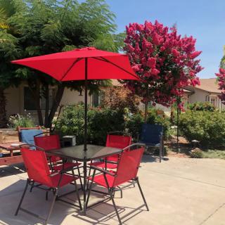 A sunny patio with a red umbrella and matching chairs, surrounded by lush greenery and flowering trees.