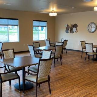 A spacious dining area with multiple tables and chairs on wooden flooring, featuring decorative wall art, a clock, and ample natural light from the windows.