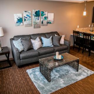A cozy living room with a gray sofa, decorative pillows, and modern artwork above. A coffee table sits on a patterned rug, and a dining area is visible in the background.