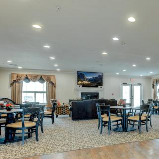 A spacious interior with a combined living and dining area, featuring several tables and chairs, a television mounted on the wall, and large windows providing natural light.