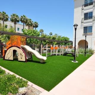 A vibrant playground with a slide and artificial grass, surrounded by palm trees and a sunny sky.