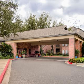 A brick building entrance with a covered driveway surrounded by trees and shrubs on a cloudy day.