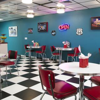 A retro-style diner with checkered flooring and red chairs, featuring a neon 'open' sign on the wall.