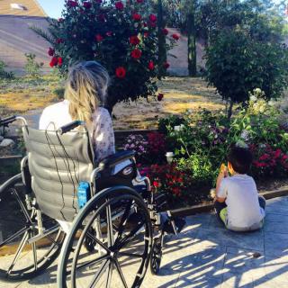 A person in a wheelchair and a child enjoying time together in a garden surrounded by flowers.