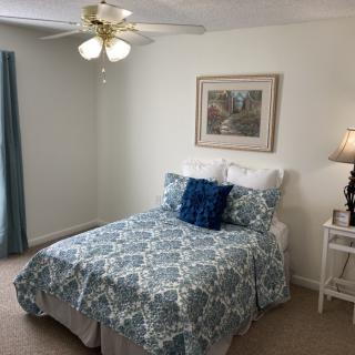 A cozy bedroom with a neatly made bed, featuring blue and white patterned bedding and a decorative pillow. A lamp and artwork add to the room's charm, while light from a ceiling fan enhances the space.