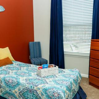 A cozy bedroom with colorful decor, featuring a bed, chair, dresser, and a large window with curtains.