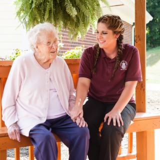 An elderly woman sitting with a caregiver on a bench, sharing a moment of companionship outdoors.