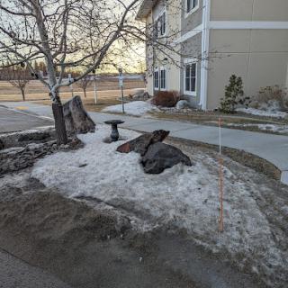 A winter scene featuring a small landscaped area with snow, rocks, and a tree beside a building.