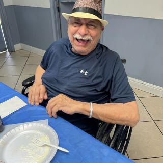 A cheerful man in a hat smiling while sitting at a table indoors.