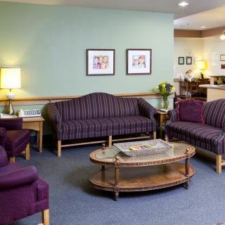 A cozy waiting room with purple sofas and chairs, a glass coffee table, soft lighting, and framed artwork on the walls.
