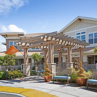 A beautifully landscaped entrance with a wooden pergola and seating area in front of a modern building.