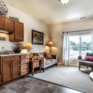 A cozy living room area with a kitchenette, featuring warm lighting and natural light from large windows.