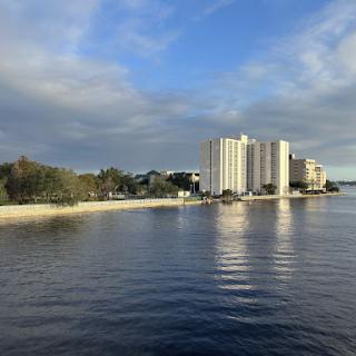 A serene view of a waterfront with modern buildings against a backdrop of clouds and clear sky.