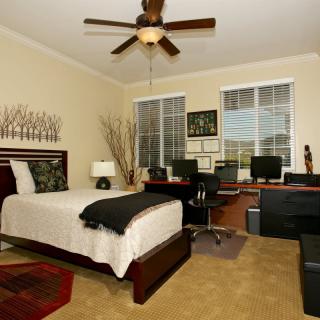 A cozy bedroom with a workspace featuring two computers, a bed, and ceiling fan.