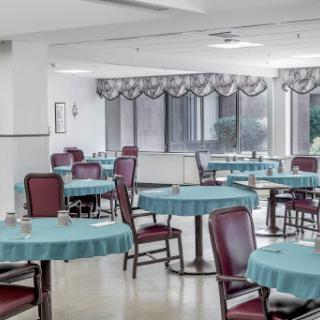 An empty dining area featuring tables with blue tablecloths and red chairs, illuminated by natural light from large windows.