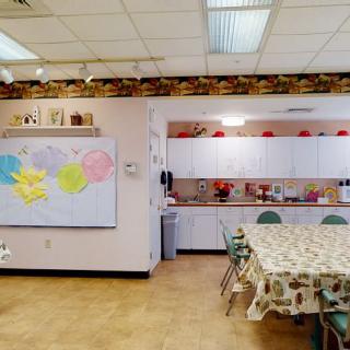 A classroom with a kitchen area, featuring a whiteboard, tables, chairs, and cabinets.