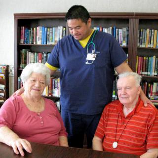 A caregiver supports two elderly individuals in a library setting.