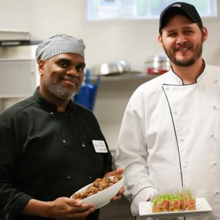 Two chefs proudly display their culinary creations in a professional kitchen.