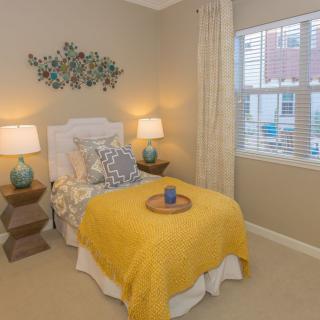 A cozy bedroom with a single bed featuring a yellow blanket and decorative pillows, flanked by two lamps on geometric side tables.
