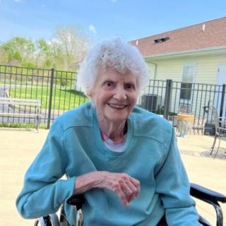 An elderly woman smiling while sitting outside in a wheelchair.