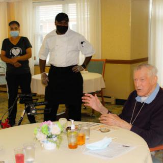 An elderly man engages in conversation at a dining table with staff members present.