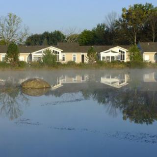 A serene lakeside scene with houses reflected on the water and mist hovering above.