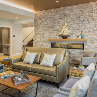 A modern living room with a stone fireplace, stylish furniture, and a wooden model sailboat on the mantel.