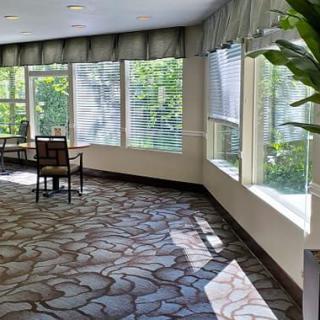 A sunlit lobby area with large windows, a table and chairs, and decorative plants.