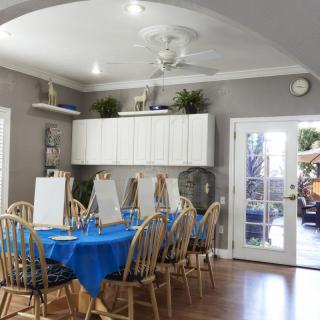 A well-lit dining area converted into an art station with easels set up for painting on a large table covered with a blue tablecloth.