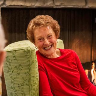 A woman in a red shirt smiling while sitting indoors near a fireplace.