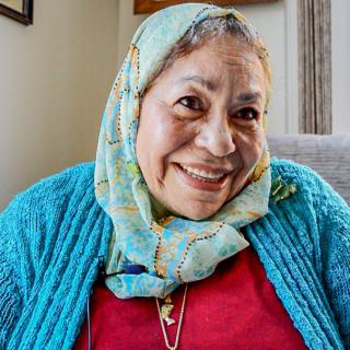 A smiling woman wearing a headscarf and a blue sweater, sitting indoors.