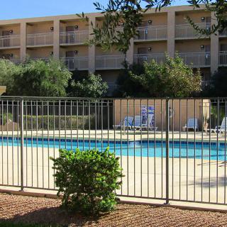 Outdoor pool area of a hotel with surrounding fence and nearby trees.