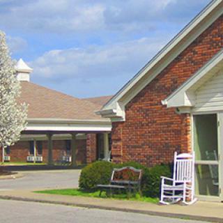 A brick building with a white rocking chair and a bench outside, next to a blooming tree.