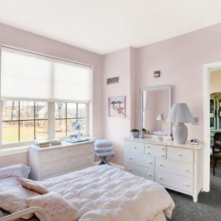 A cozy bedroom with pastel walls, featuring white furniture and a large window allowing natural light to fill the room.