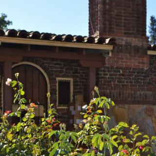 A rustic brick building with a chimney, surrounded by lush garden plants and an arched doorway.