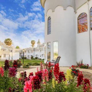 A picturesque garden with vibrant flowers in front of a white building under a partly cloudy blue sky.