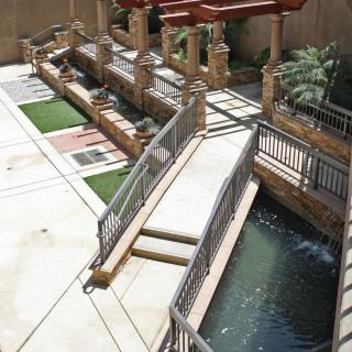 A scenic outdoor area with a wooden bridge over a water feature, surrounded by landscaped gardens and architectural elements.