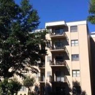 A multi-story apartment building with balconies, surrounded by trees under a clear blue sky.