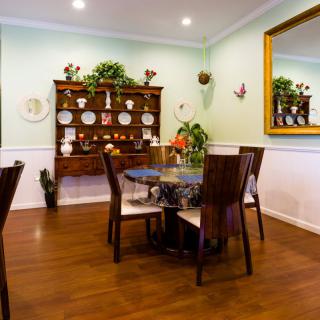 A cozy dining room with a circular table and wooden chairs, decorated with a mirror and a shelf displaying plates.