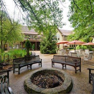 A tranquil outdoor garden area featuring benches around a circular fire pit, surrounded by lush trees and patio umbrellas.