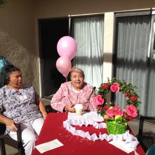 An elderly woman celebrating her birthday outdoors with balloons and flowers, accompanied by another person.