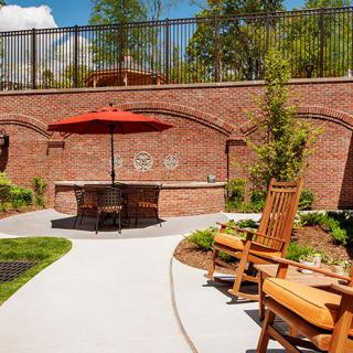 A cozy patio area with wooden chairs, a red umbrella, and a brick wall backdrop surrounded by lush greenery.