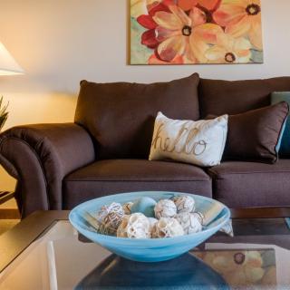 A cozy living room with a brown sofa, decorative cushions, and a glass coffee table featuring a blue bowl with ornamental balls. A colorful painting and lamp enhance the warm ambiance.