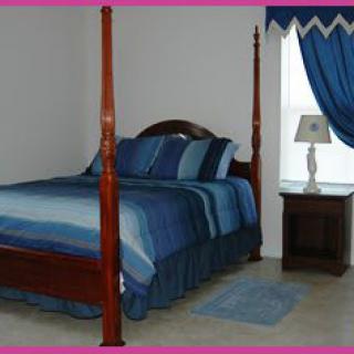 A classic bedroom with a four-poster bed adorned with blue bedding, next to a nightstand and lamp under matching blue curtains.