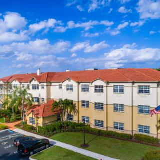 Aerial view of a residential apartment building with a red roof, surrounded by palm trees and a parking area under a partly cloudy sky.