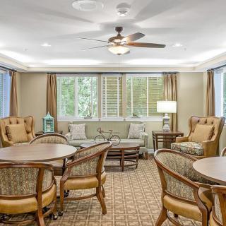 A cozy living room with comfortable seating arrangements, including sofas and chairs around tables, complemented by a ceiling fan and large windows.
